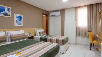 A hotel room with two beds, a desk, a chair, an air conditioner, and two framed pictures on the wall.