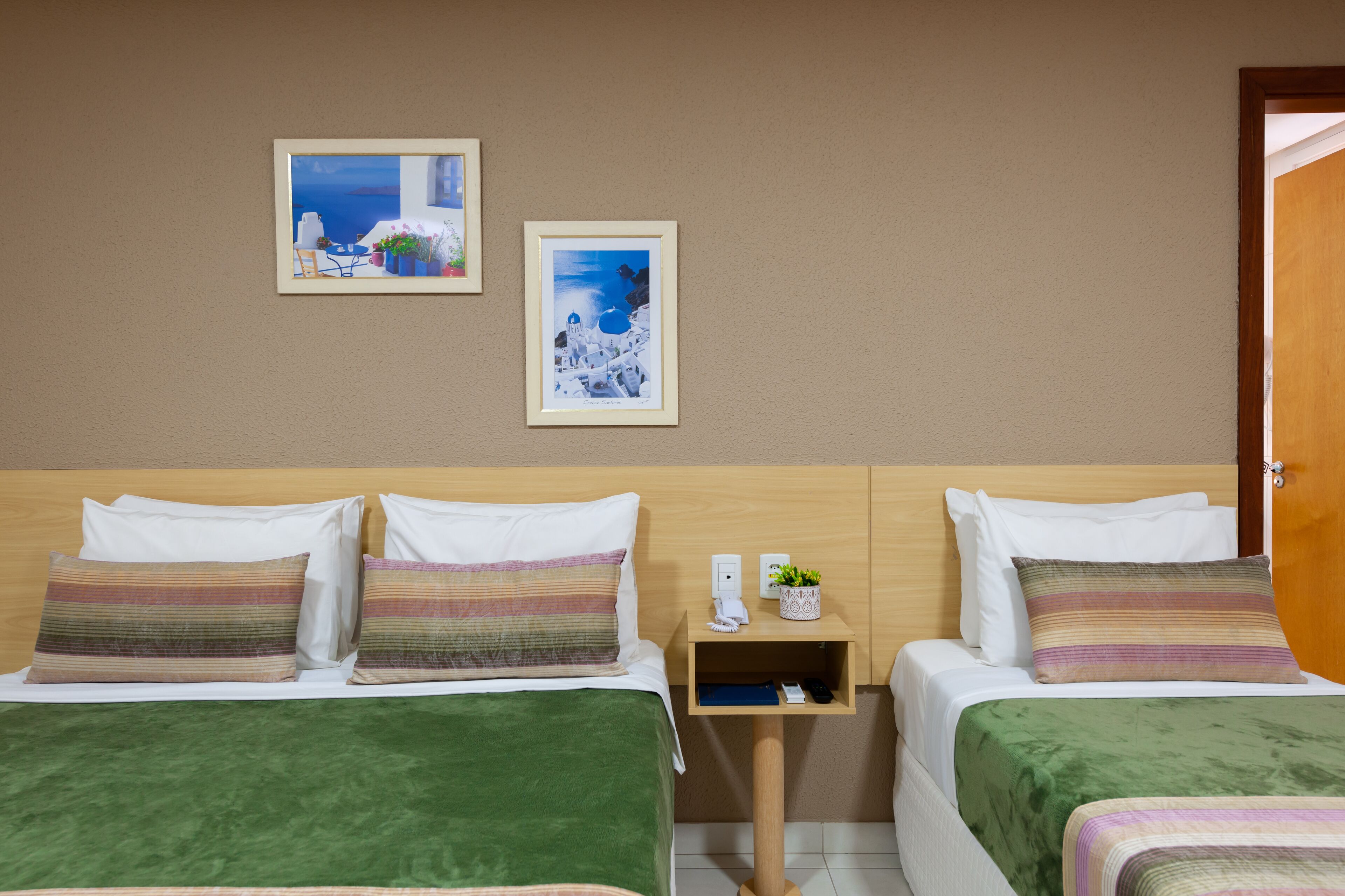Habitación de hotel con dos camas, cabecera de madera, mesita de noche con un teléfono y una planta pequeña, y dos cuadros en la pared.