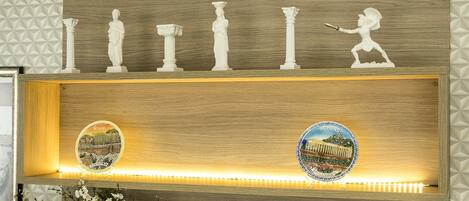 Un mostrador de recepción con detalles de madera, estatuas decorativas blancas y carteles iluminados que muestran puntos emblemáticos de Atenas.