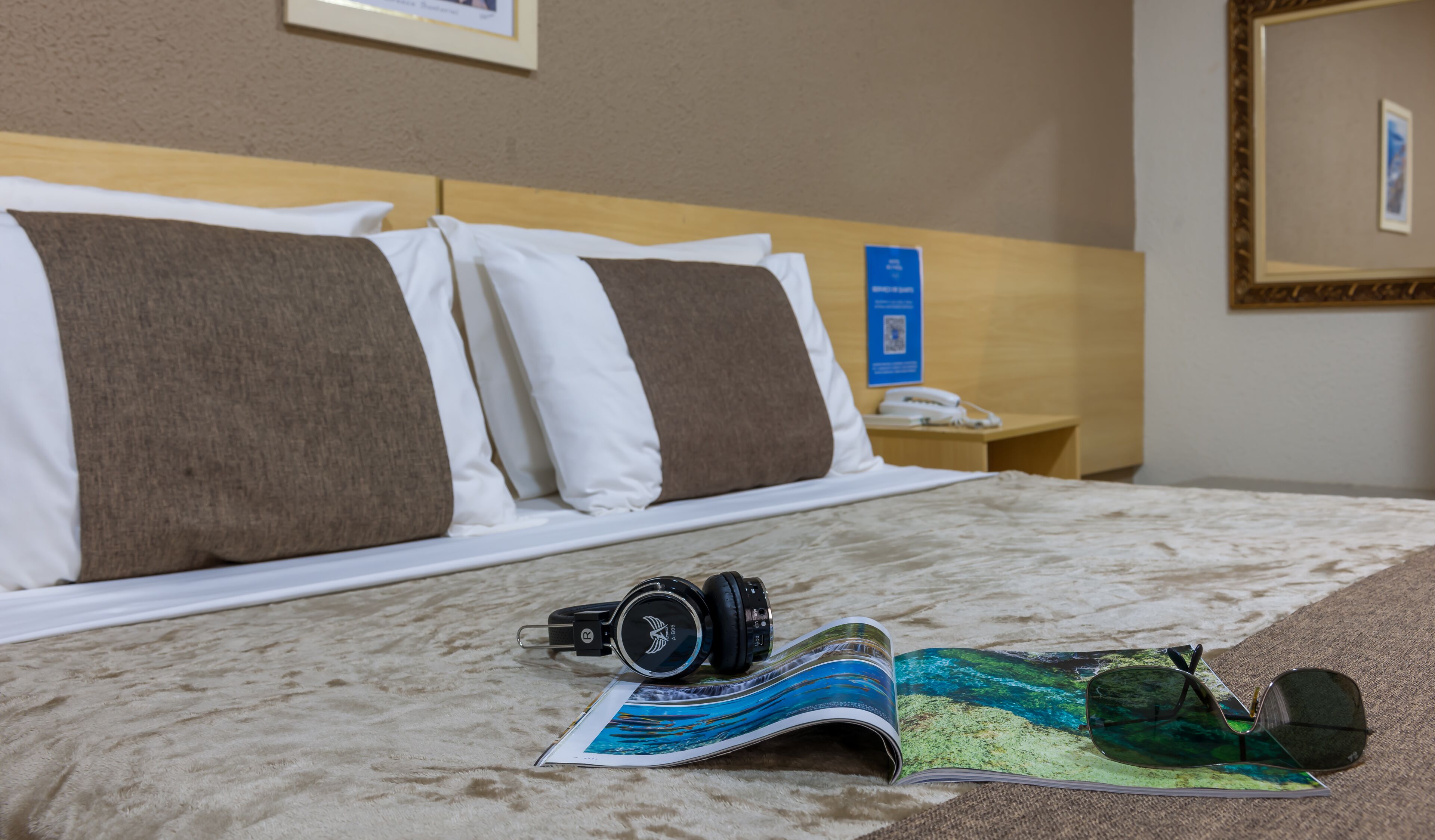A neatly made bed with pillows, a pair of headphones, and a magazine on a carpeted floor.