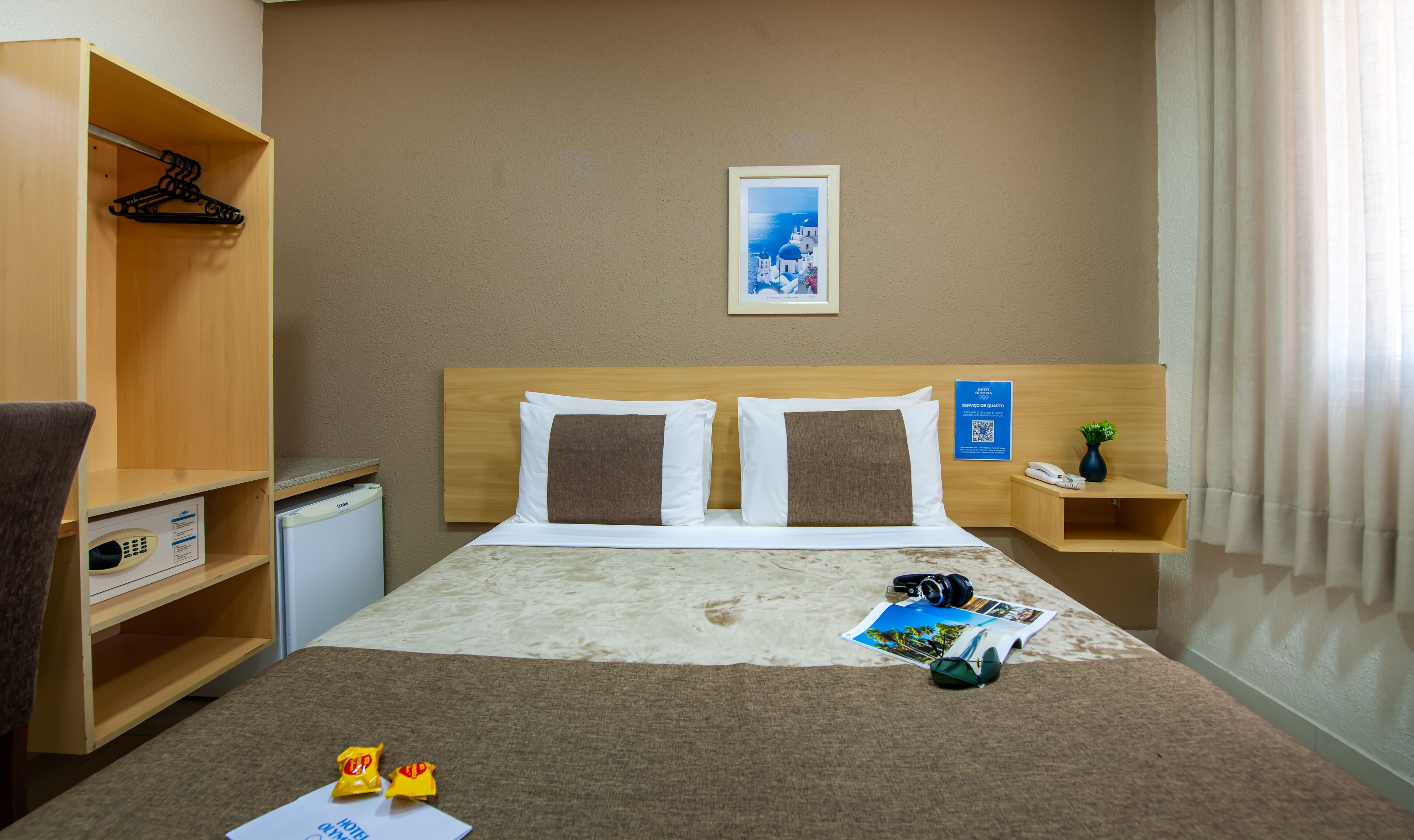 A hotel room with a bed, a wooden headboard, a small desk, a chair, a minibar, and a wall-mounted phone.