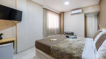 A hotel room with a bed, a TV, a small desk with a plant, a suitcase, and an air conditioner.