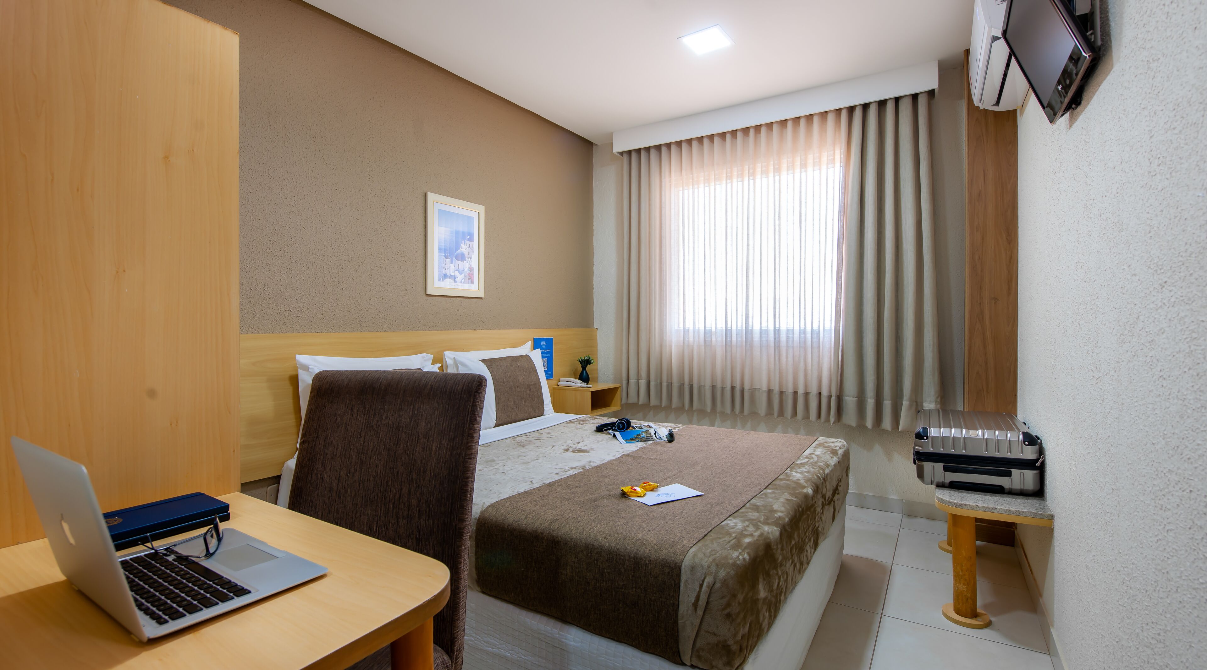 Habitación de hotel con cama, escritorio con ordenador portátil, silla, mesita de noche con teléfono y maleta.