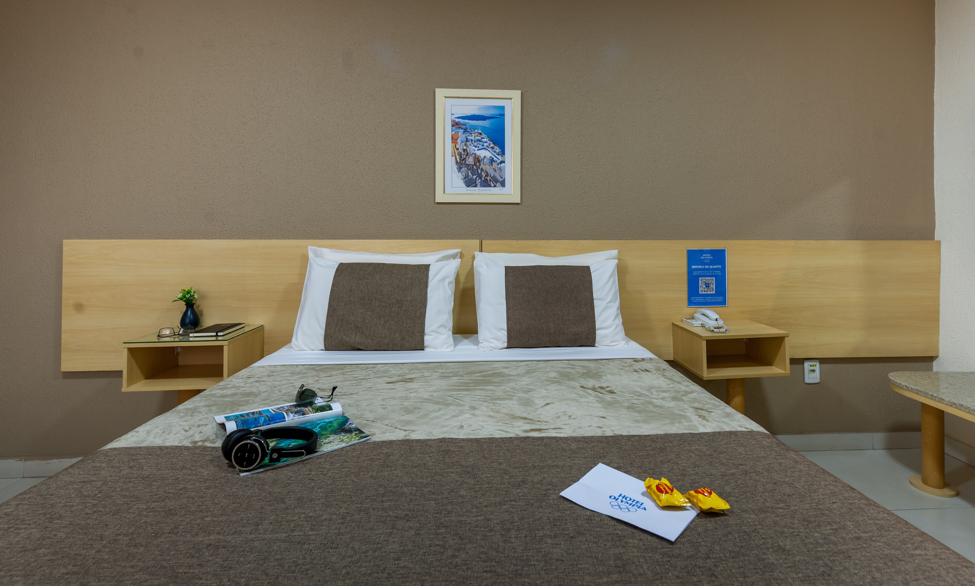 Quarto de hotel com cama, duas mesas de cabeceira, um quadro emoldurado, uma planta pequena, um par de fones de ouvido e um guia de boas-vindas.