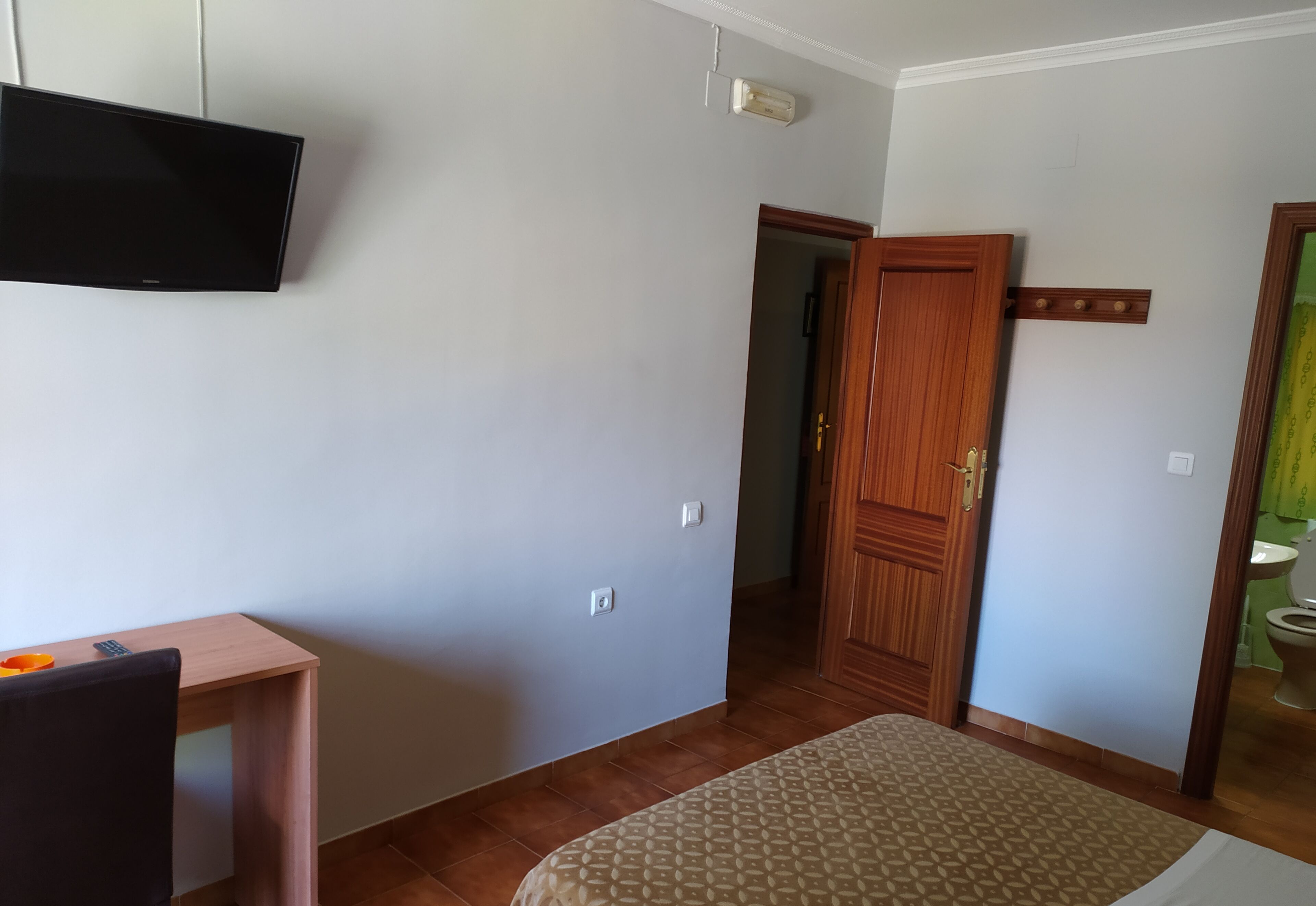 Una habitación con una cama, un televisor montado en la pared, una puerta de madera y una mesita con un control remoto.