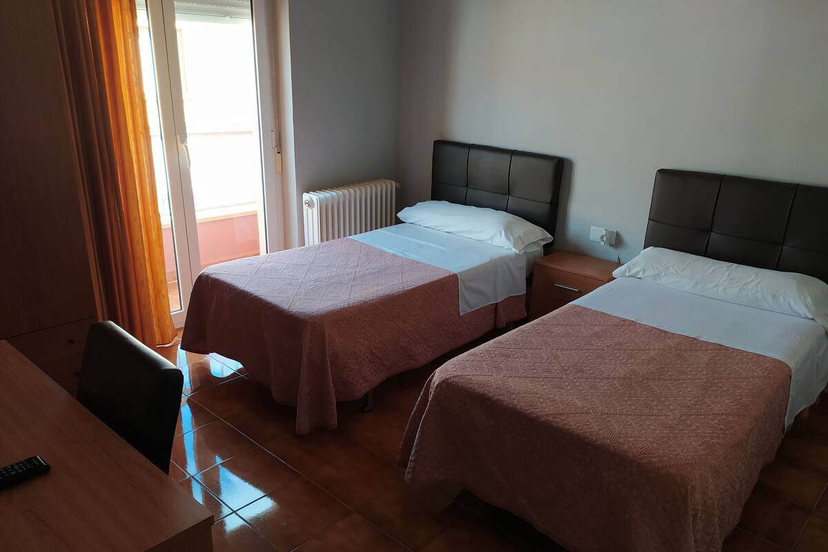 Quarto de hotel com duas camas, uma escrivaninha de madeira e uma janela com cortinas.