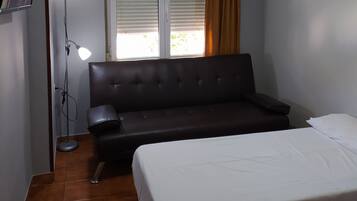 A bedroom with a bed, a sofa, a ceiling fan, a window with blinds, a floor lamp, and a TV on the wall.