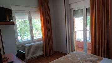 A bedroom with a bed, a desk, a chair, a TV, and a window with curtains.