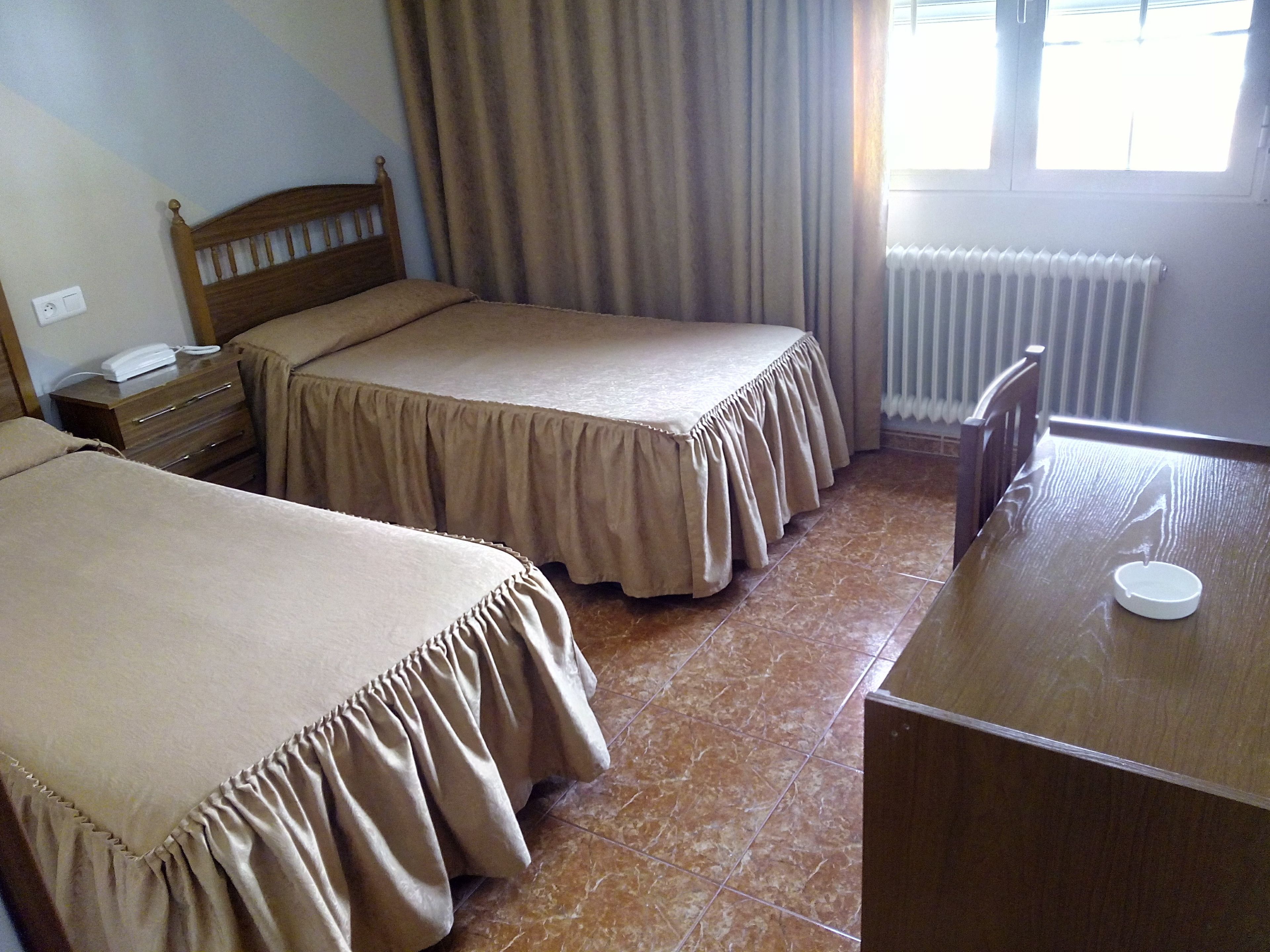 Una camera d'albergo con due letti singoli, un comodino in legno con il telefono, una scrivania con una sedia e un termosifone.