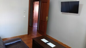 A room with a brown sofa, a wooden table with a remote control, and a TV on the wall.