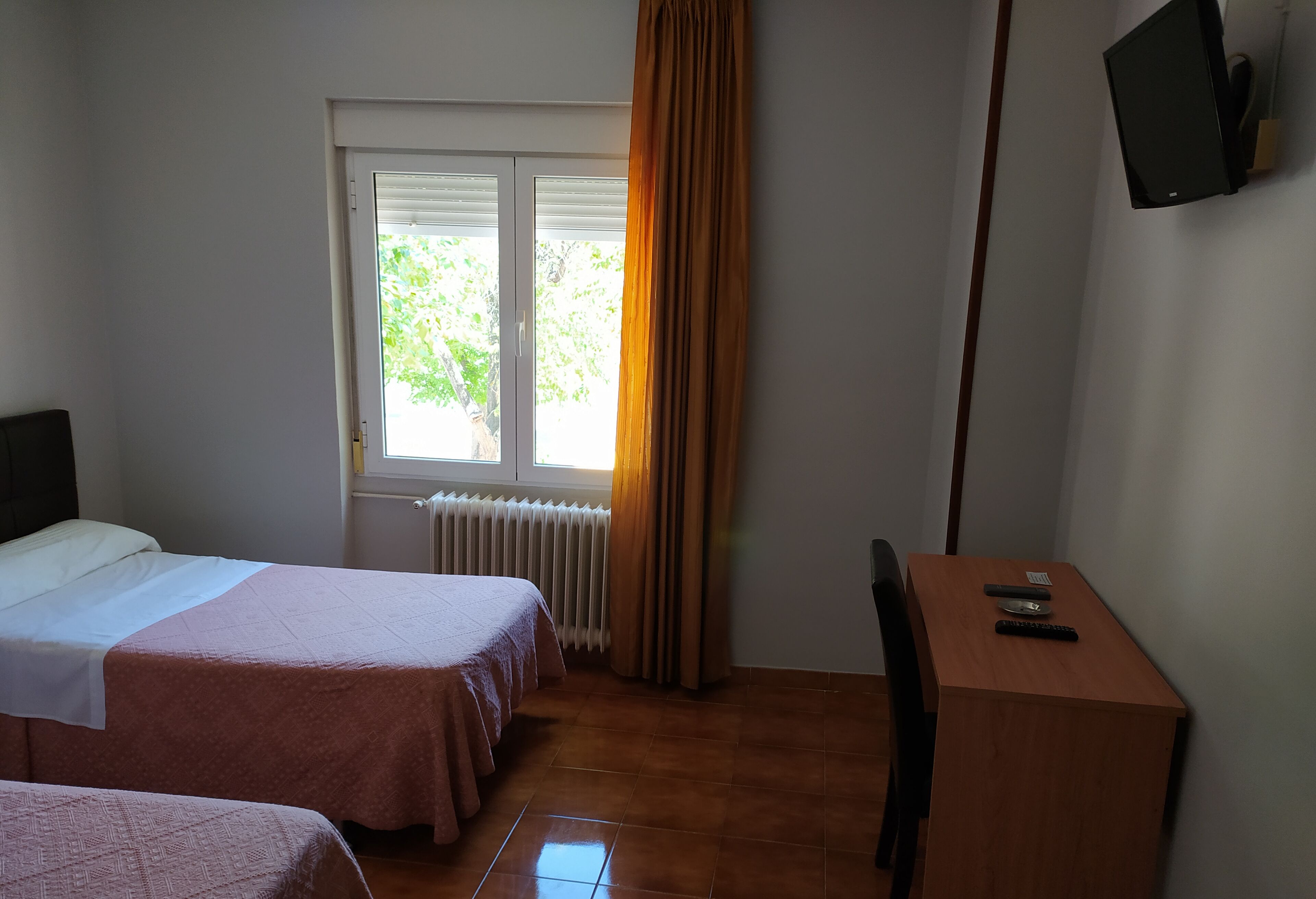 A hotel room with two beds, a desk, a chair, a TV mounted on the wall, and a window with curtains.