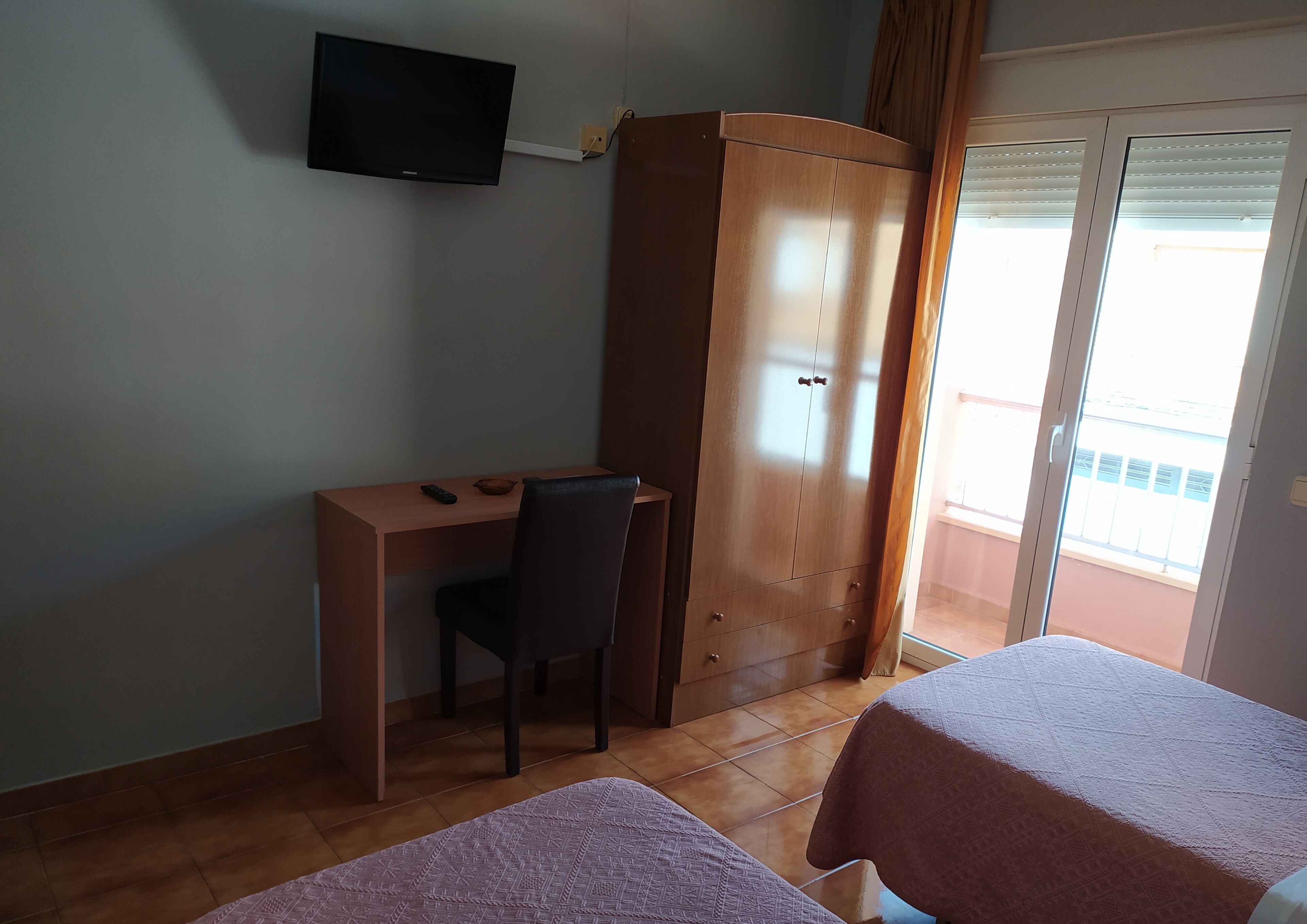 A room with a wooden desk, a chair, a wardrobe, a television on the wall, and two beds with patterned covers.