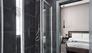 A modern bathroom with a glass-enclosed shower and a separate bedroom with a bed and bedside table. - Keb Hotel (Milan)
