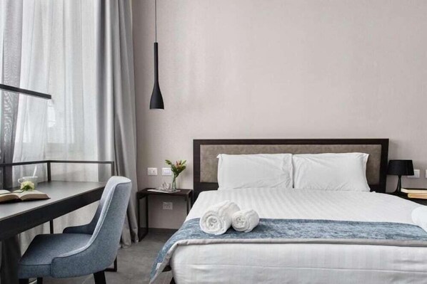 Quarto de hotel com cama, escrivaninha, cadeira, mesa de cabeceira e abajur.