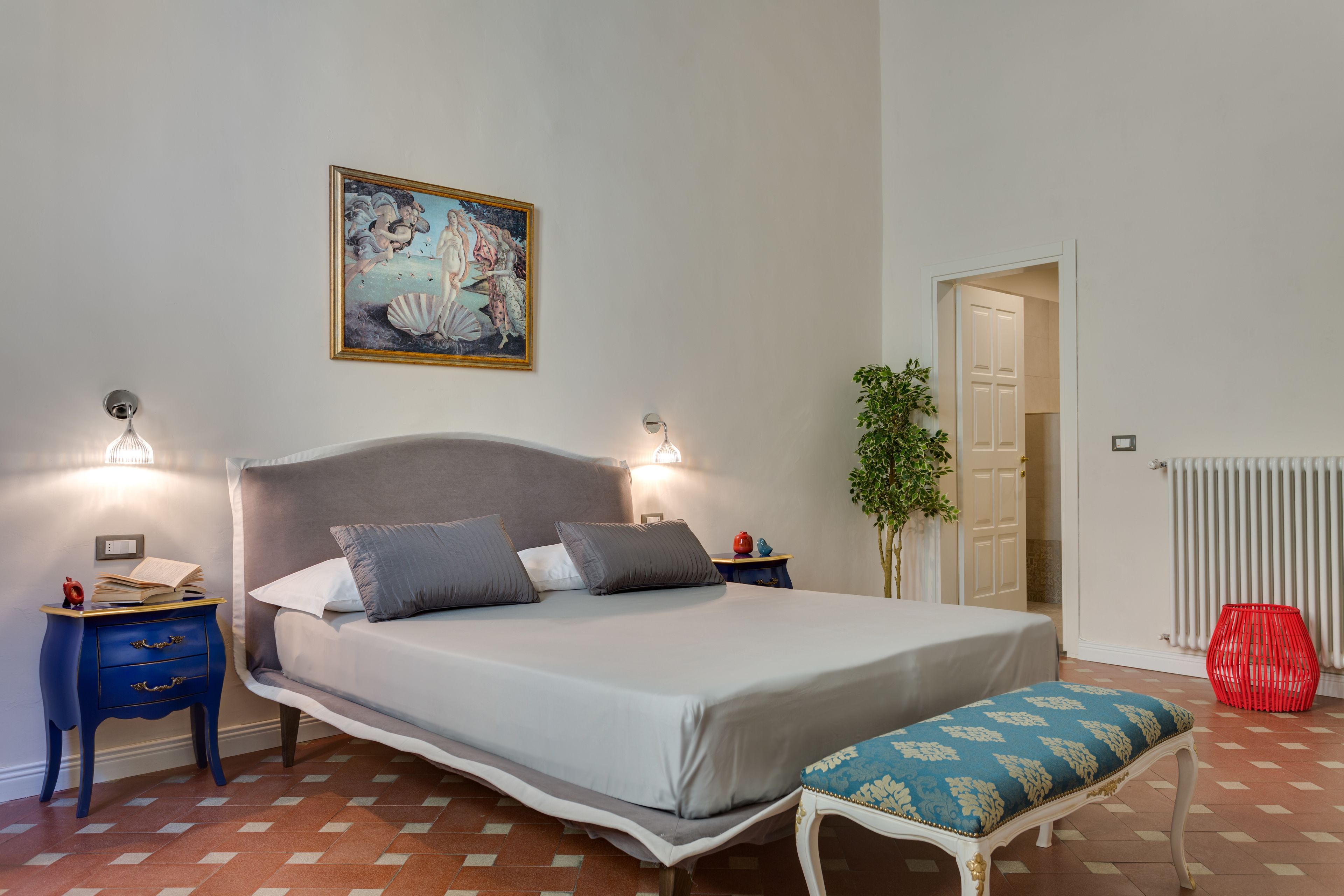 Un dormitorio con cama, mesitas de noche, banco, un cuadro, planta y radiador.