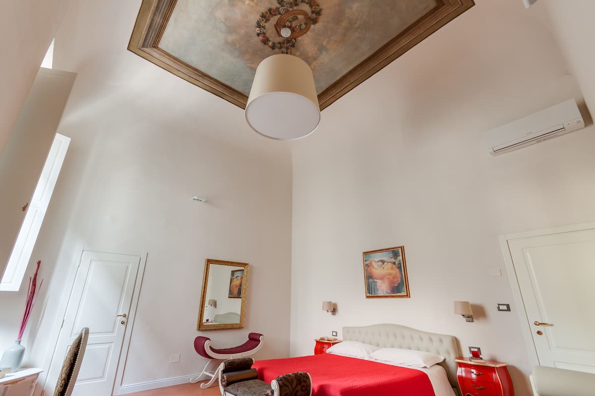 A hotel room with a large bed, a red accent wall, a ceiling with a decorative design, and a white air conditioning unit.