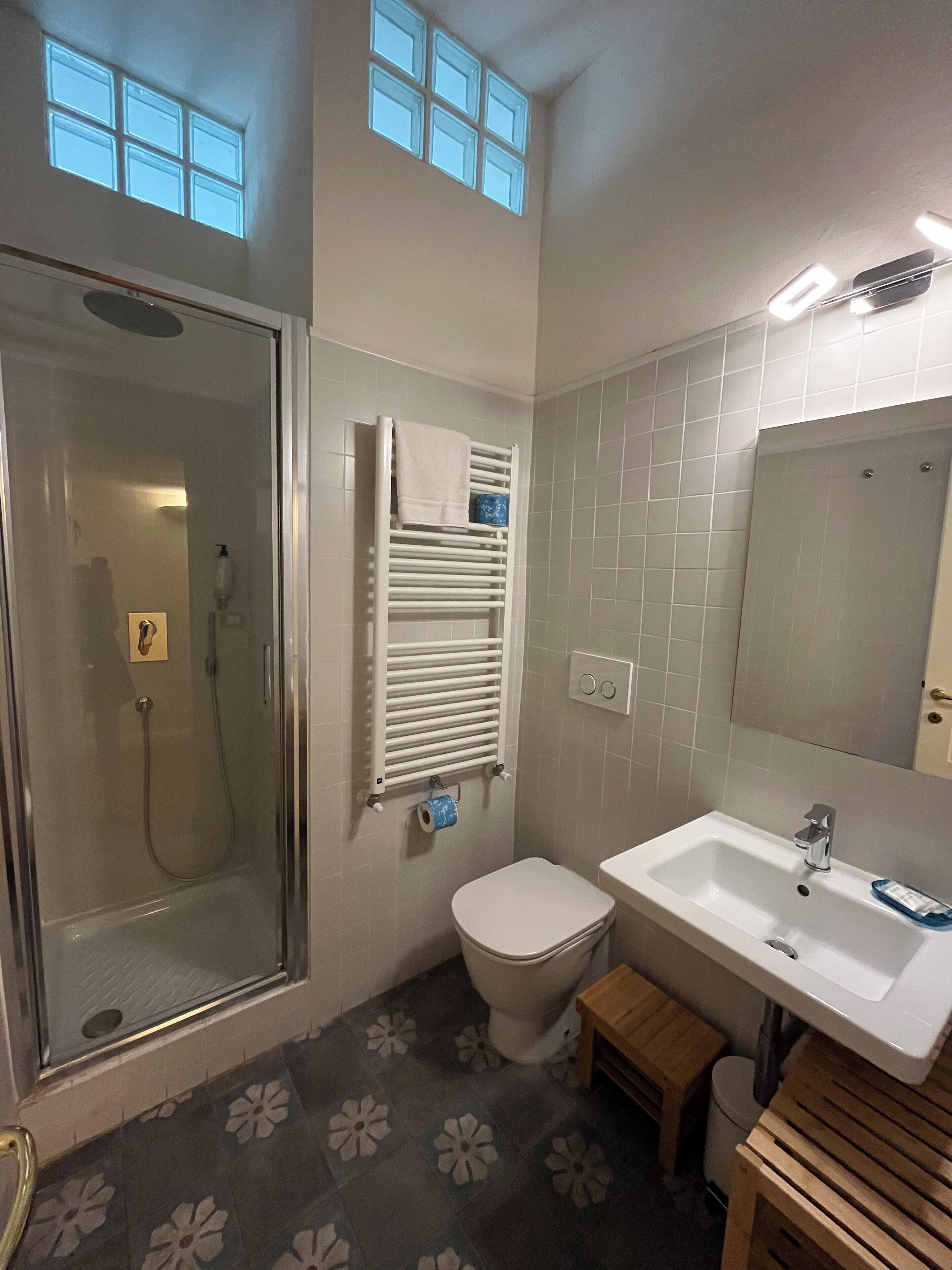 A bathroom with a glass-enclosed shower, a toilet, a sink, and a small wooden stool.