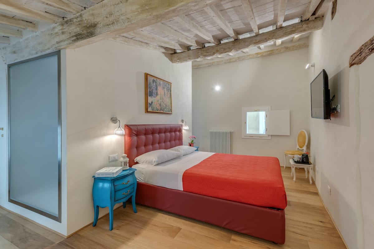A bedroom with a red bed, a blue nightstand, a white radiator, and a television on the wall.