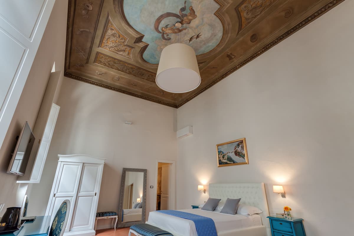 A hotel room with a large bed, a wardrobe, a mirror, a ceiling with a fresco, and wall-mounted lights.