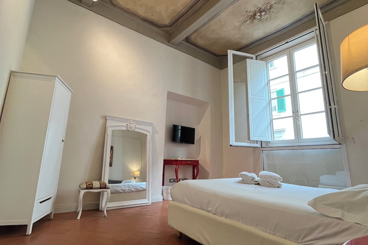 A hotel room with a large bed, a wardrobe, a vanity with a mirror, a television on the wall, and a window with a view of the outside.