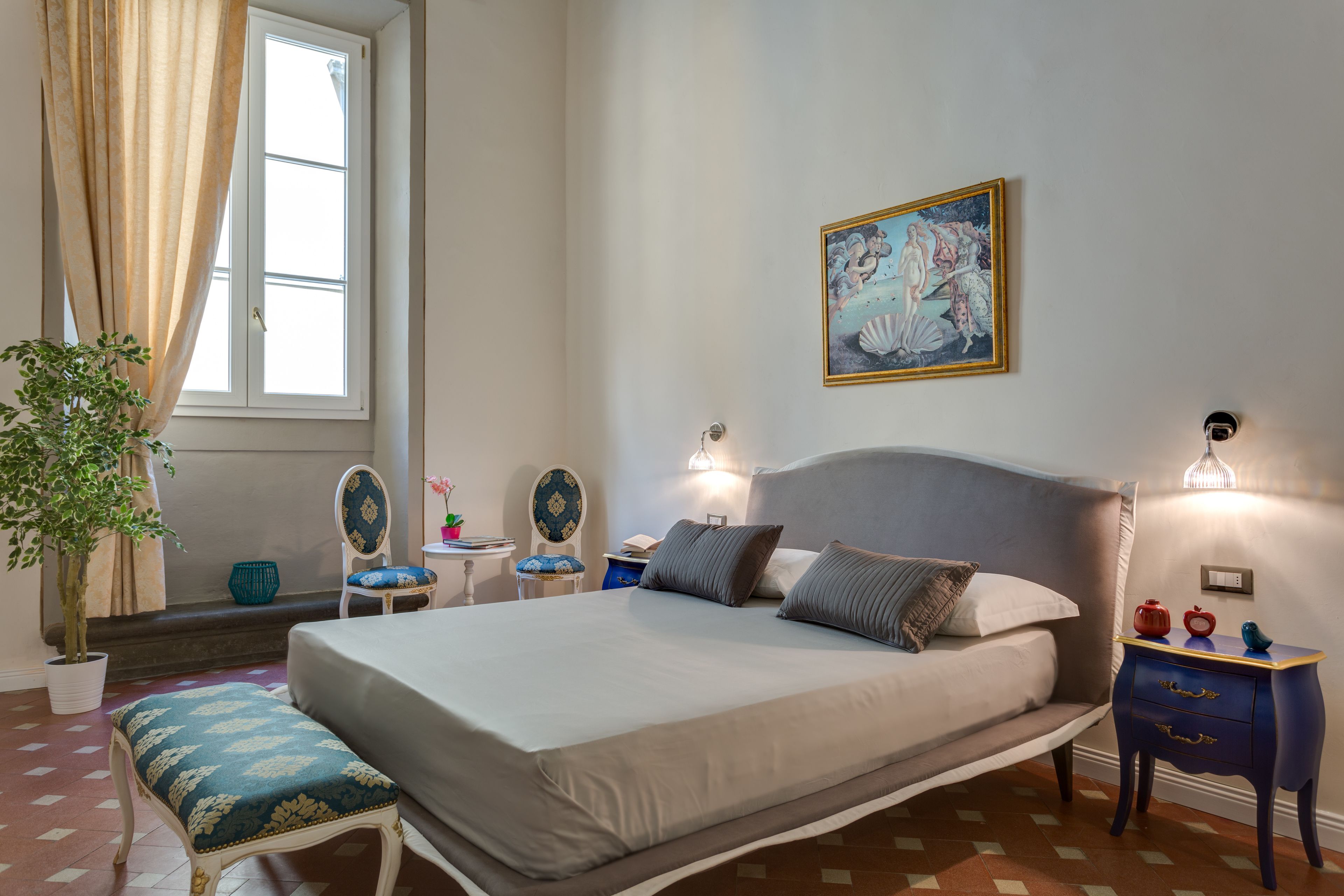A bedroom with a bed, bedside tables, a nightstand, a chair, a potted plant, and a painting on the wall.