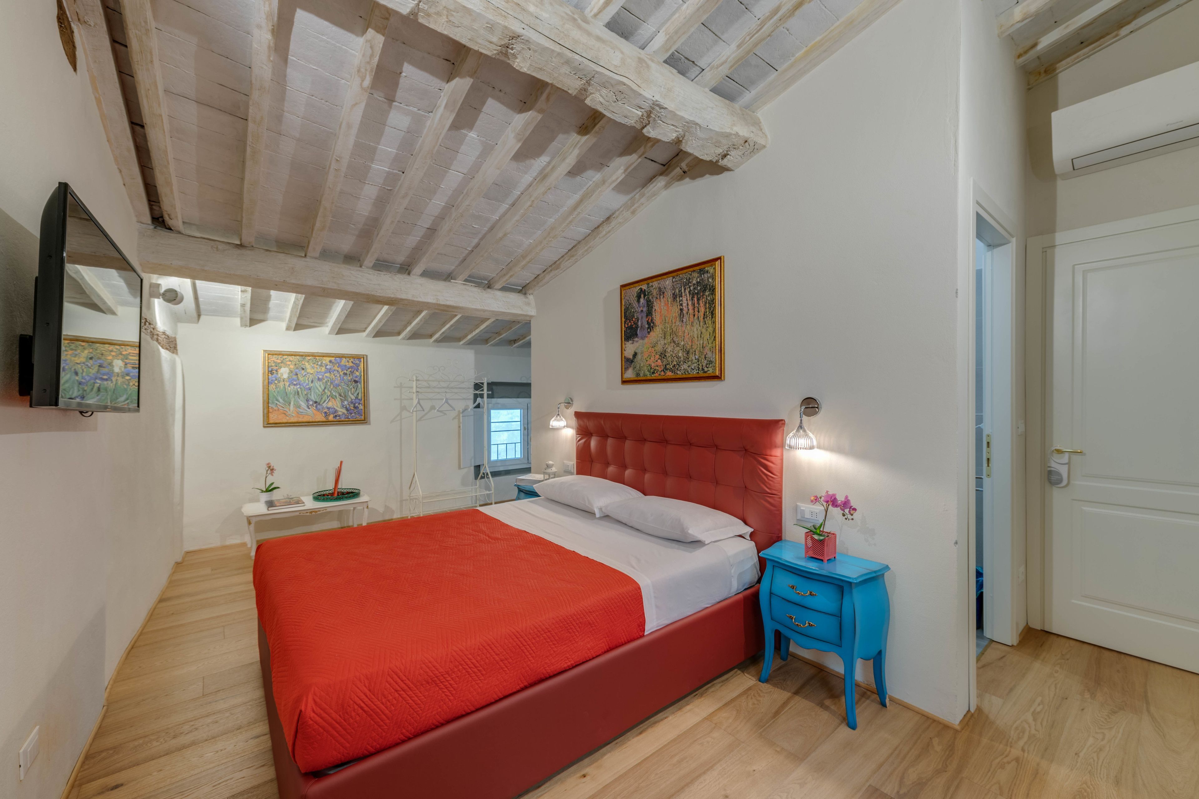 A bedroom with a red bed, a blue nightstand, and a painting on the wall.