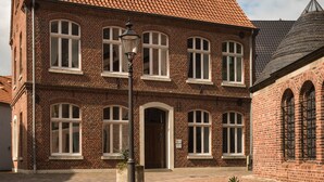A two-story brick building with a tiled roof, a central entrance, and a flower bed in the foreground. - Hotel Nagel (Sudlohn)