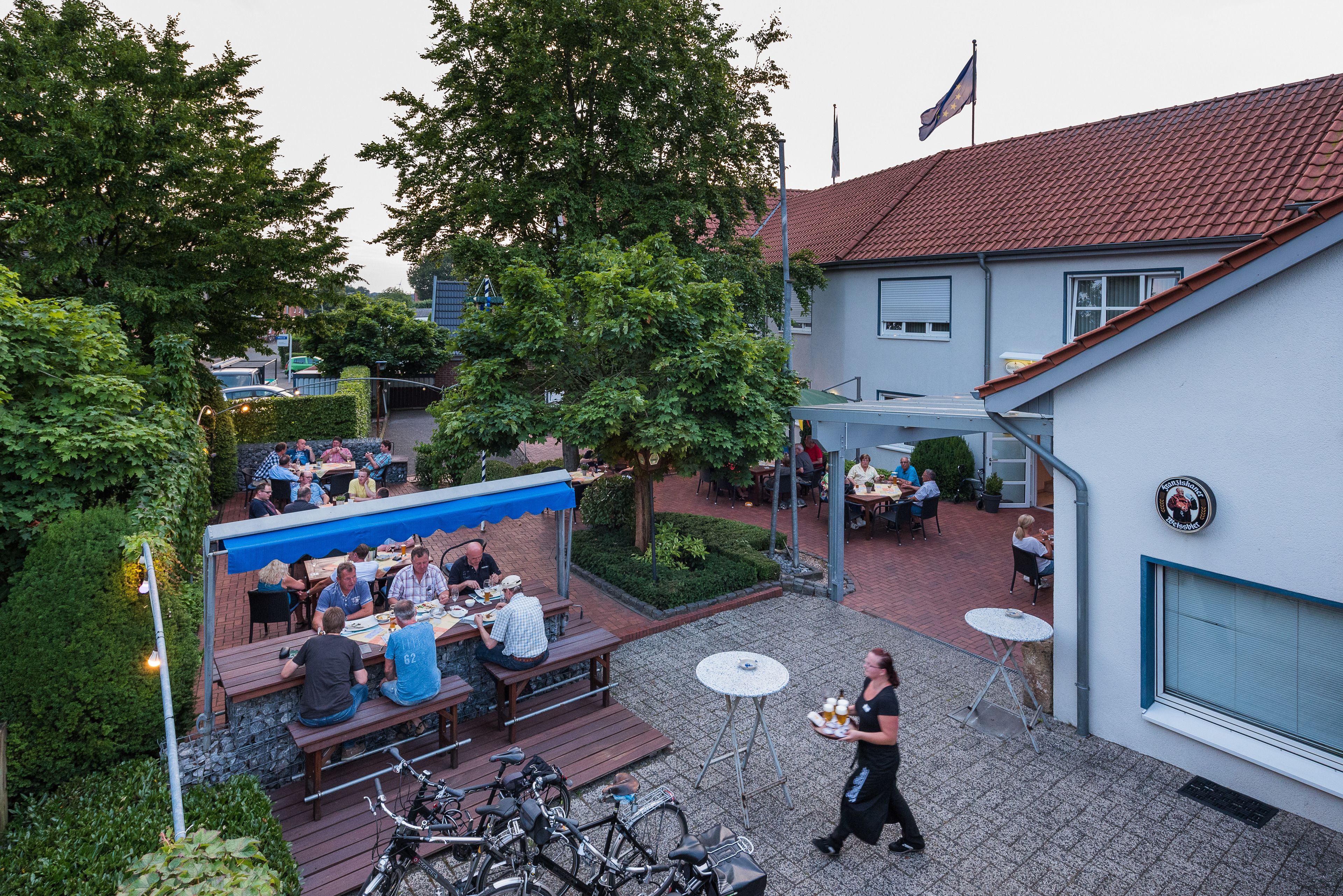 An outdoor patio area with tables and benches, surrounded by greenery and a building with a tiled roof.