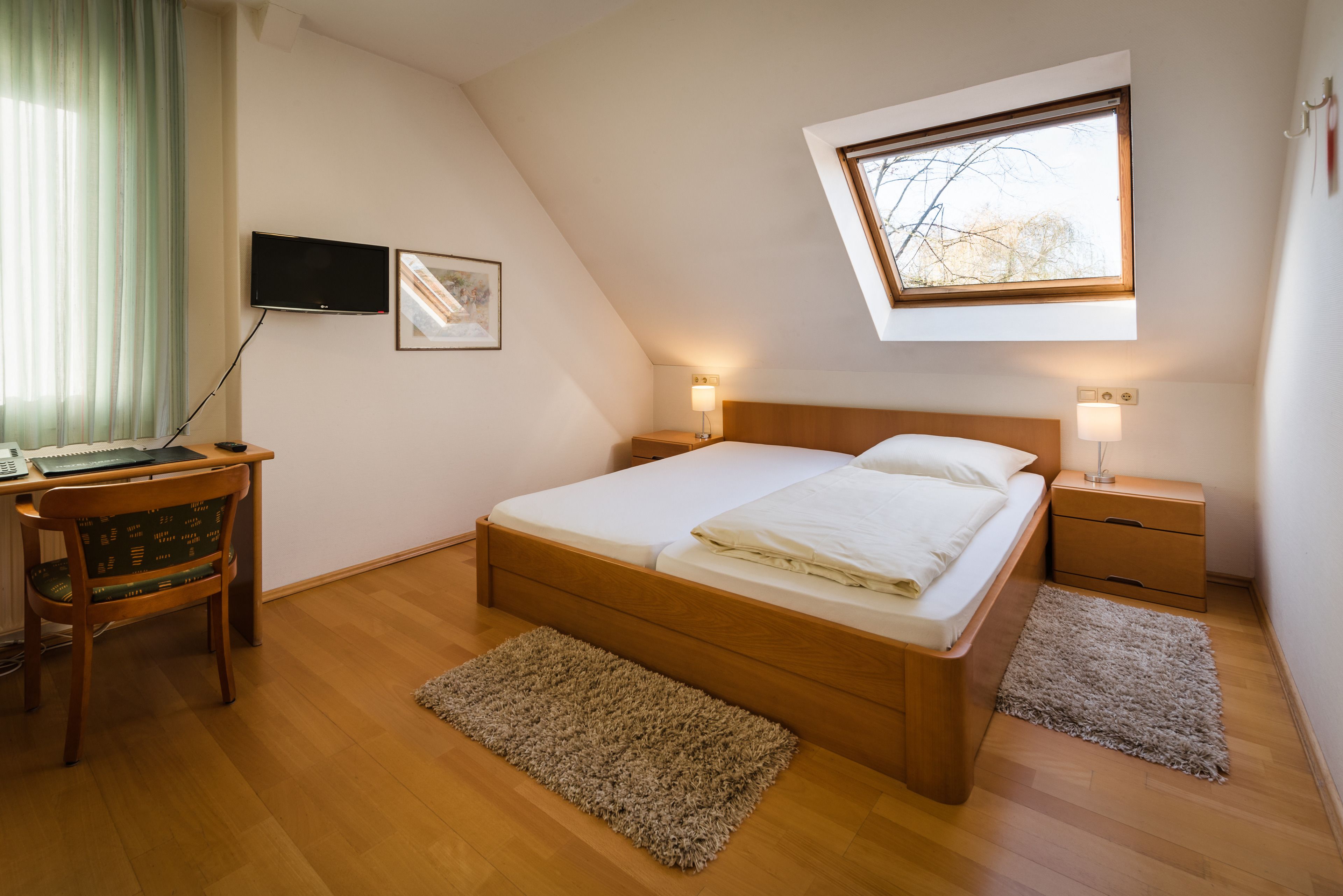 Een slaapkamer met een houten bed, een bureau met stoel, een televisie en een raam met uitzicht op bomen.