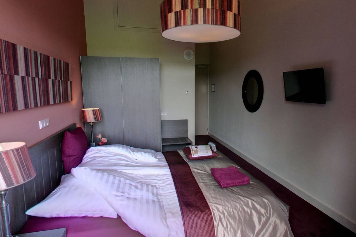 A hotel room with a bed, a bedside table, a lamp, a TV, and a wall-mounted mirror.