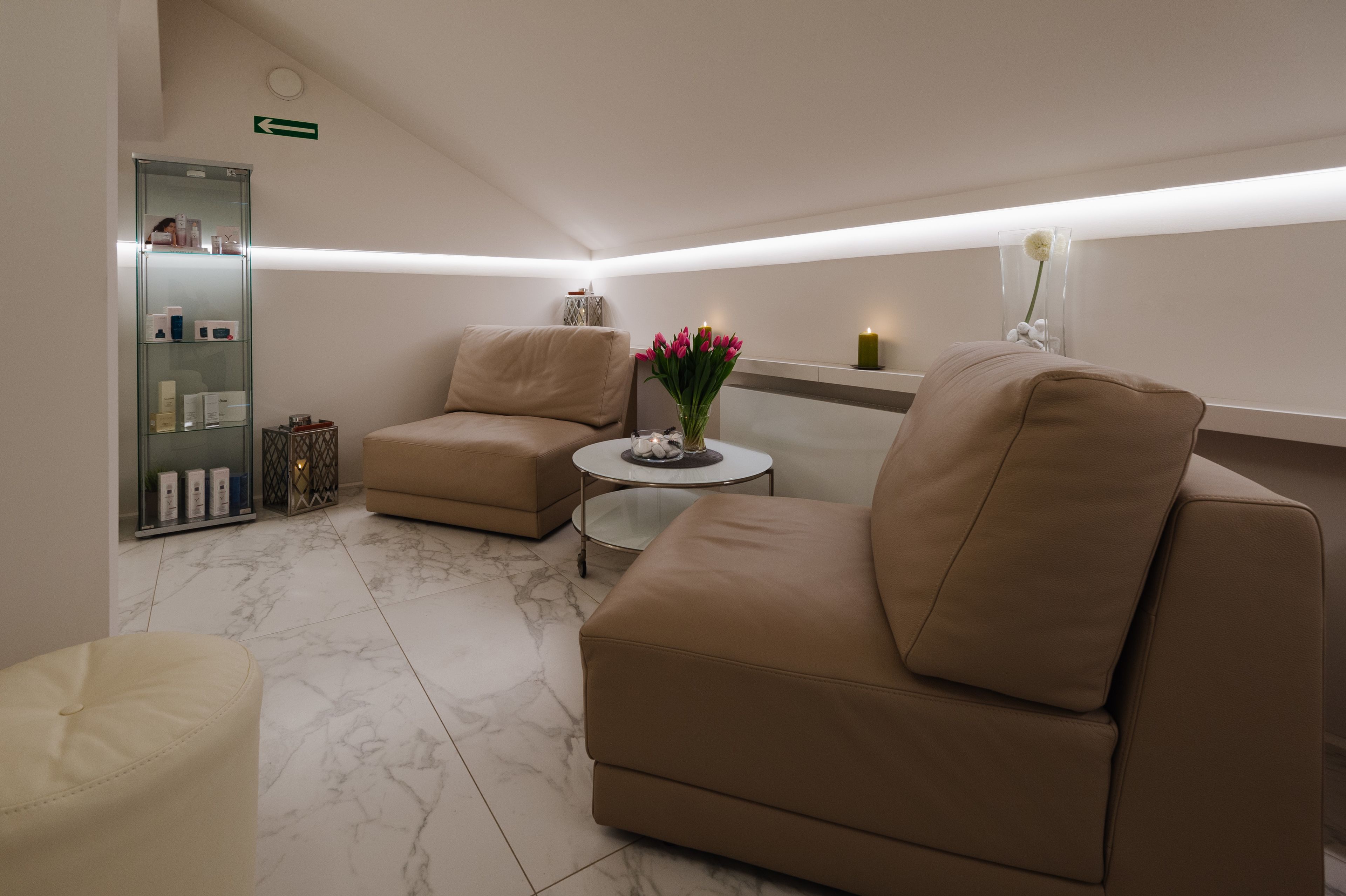 A modern, well-lit room with a beige sofa, a small round table, and a glass cabinet with toiletries.