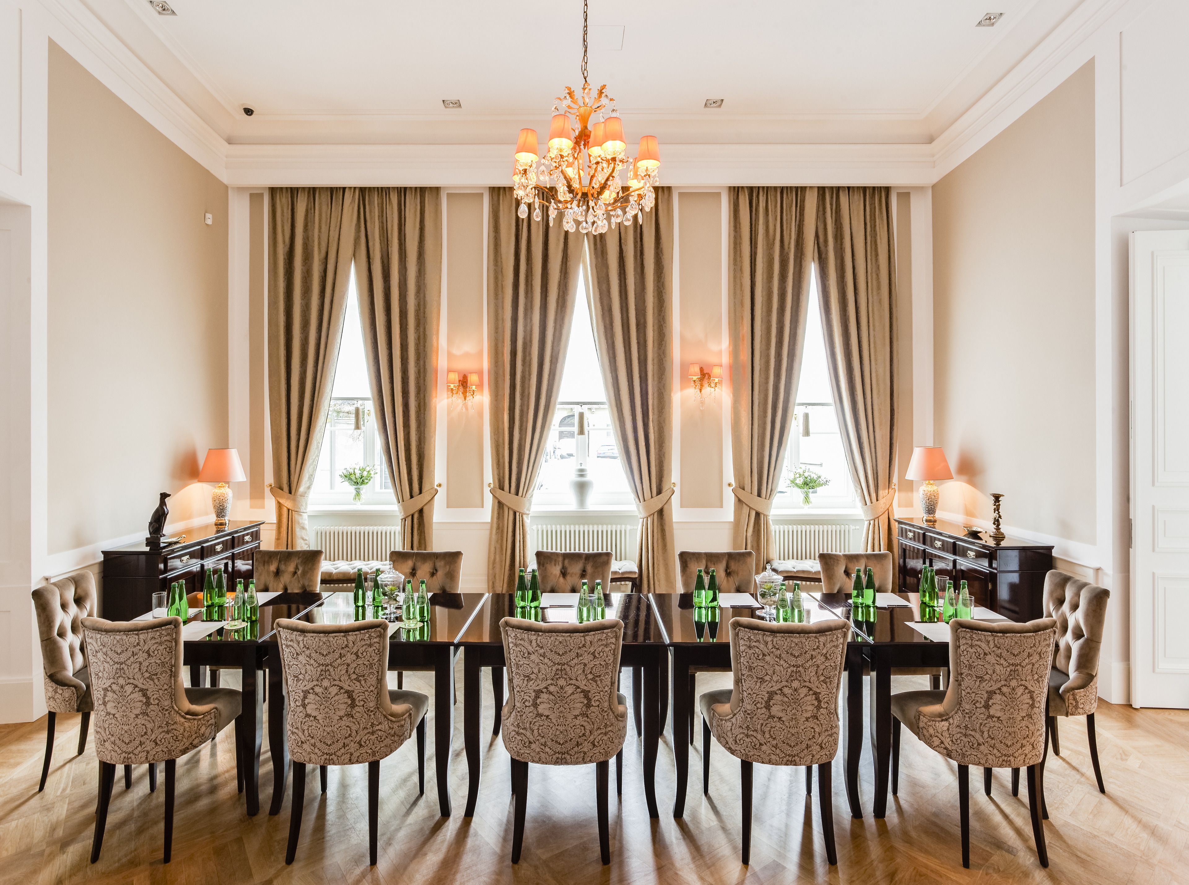 A formal dining room with a long table set for eight, flanked by tall windows with curtains, a chandelier, and wall sconces.