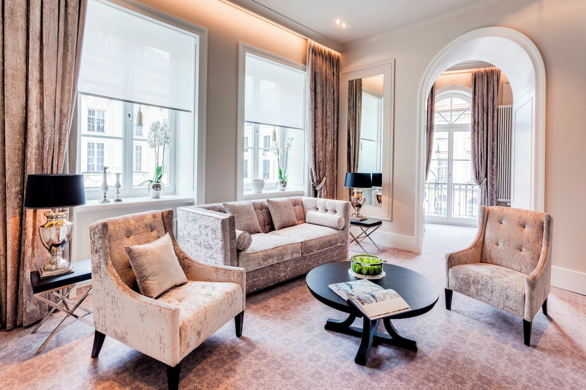 A spacious living room with a sofa set, a coffee table, and a side table with lamps. Large windows provide natural light and views of the city.