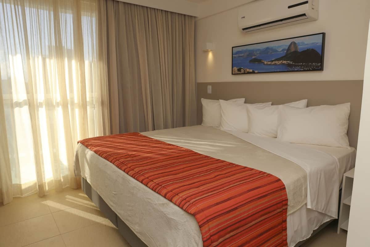 A hotel room with a large bed, a window with sheer curtains, an air conditioning unit, and a painting of a mountain landscape.