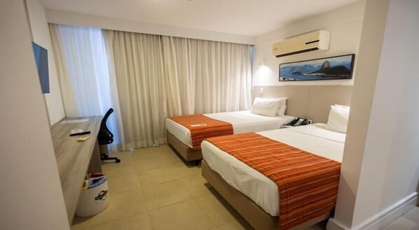 A hotel room with two beds, a desk, a chair, a trash bin, and an air conditioning unit.