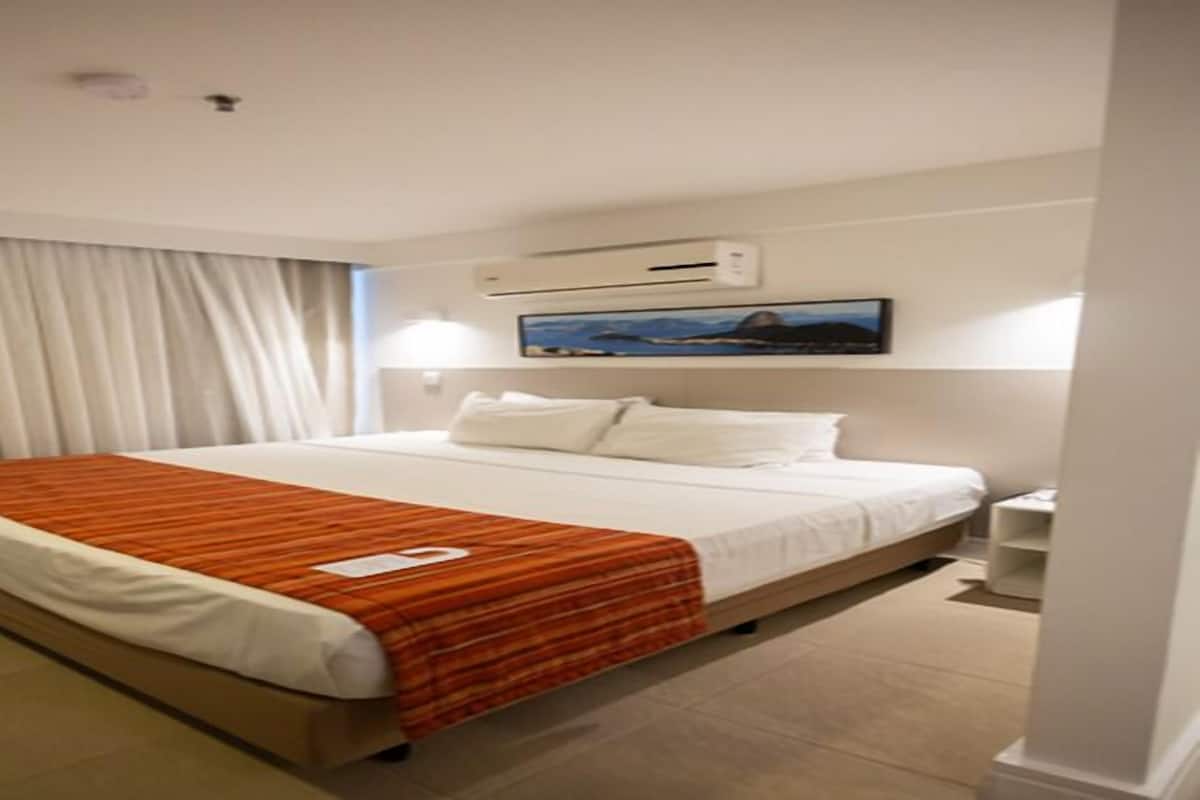 A hotel room with a large bed, white curtains, a painting on the wall, and a nightstand.