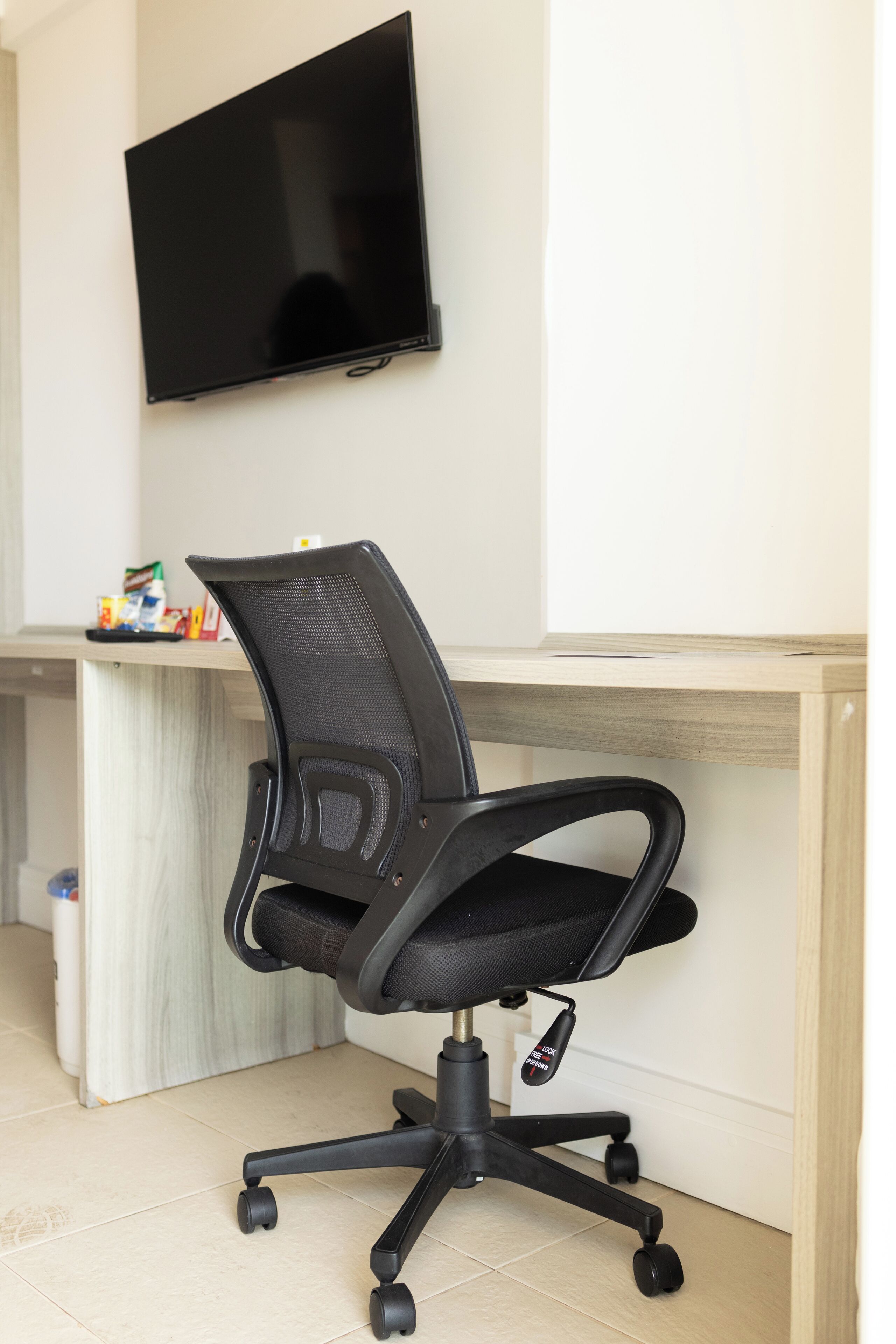Une chaise de bureau noire à roulettes, un téléviseur à écran plat fixé au mur et un bureau en bois à la finition de couleur claire.