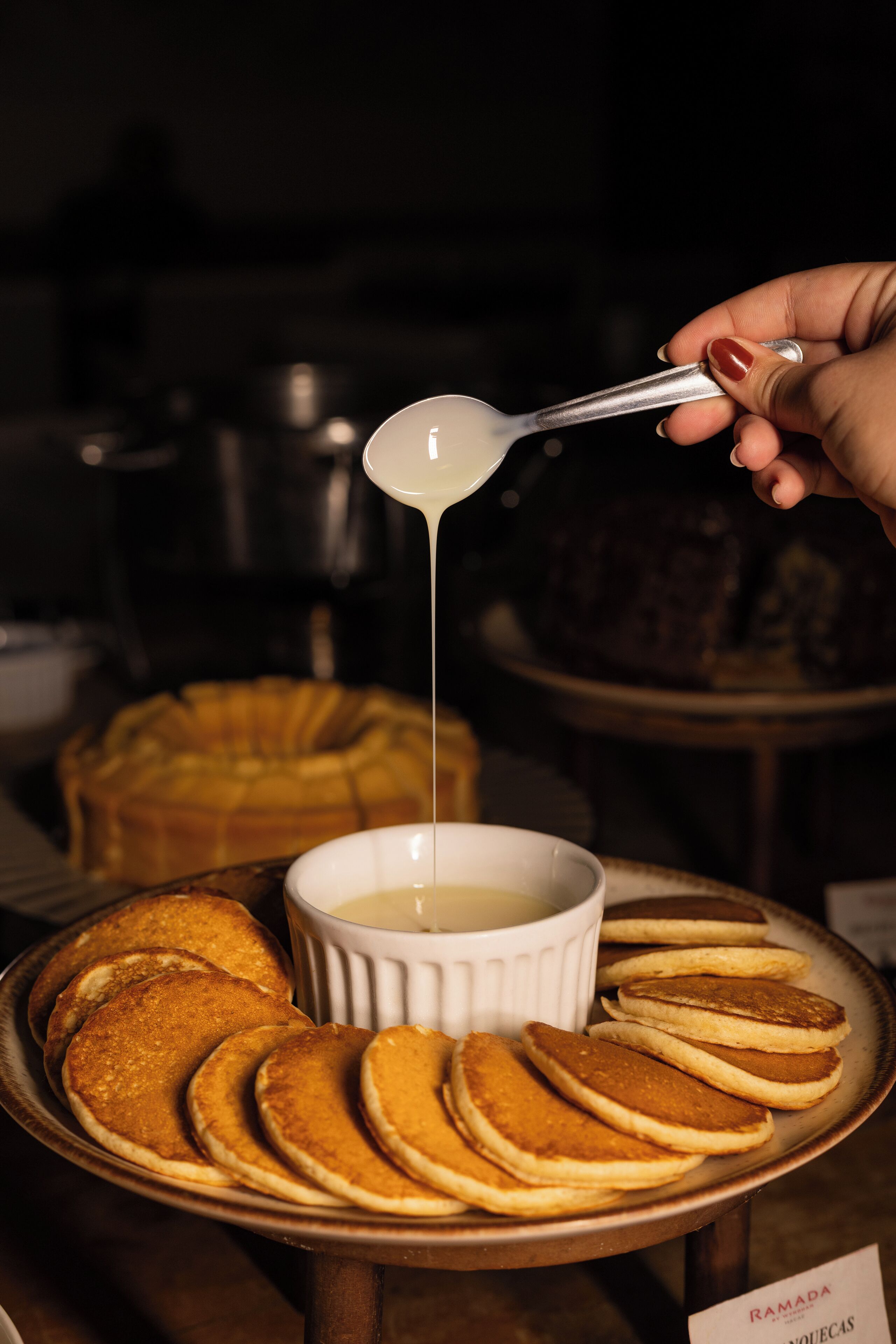 A hand pouring syrup over pancakes on a plate, with a bowl of syrup in the background.
