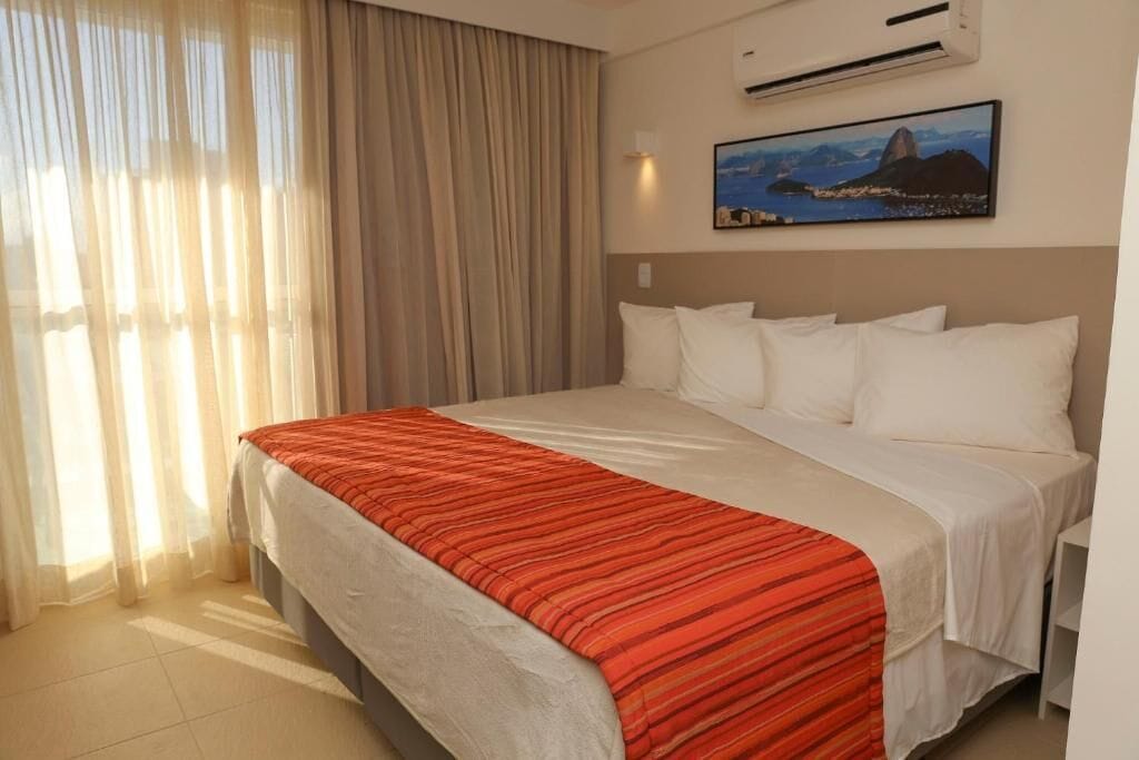 A hotel room with a large bed, beige curtains, an air conditioning unit, and a painting of a mountain landscape.