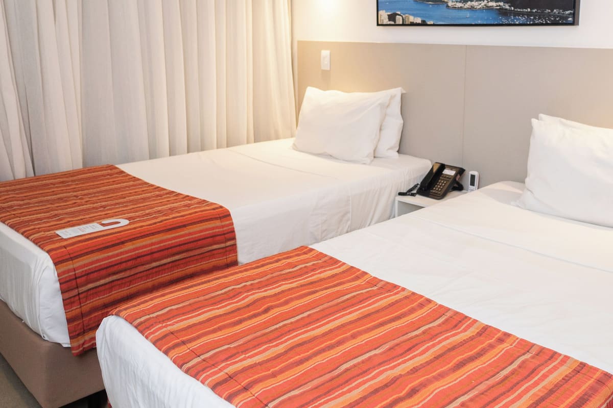 A hotel room with two beds, a painting of a coastal landscape, an air conditioning unit, and a telephone on the nightstand.