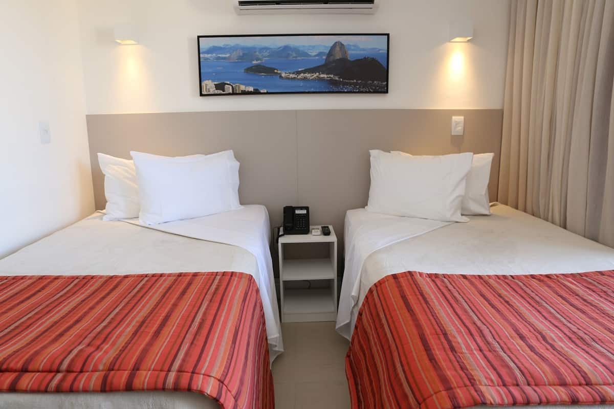 A hotel room with two beds, a small bedside table with a phone, and a wall-mounted TV displaying a cityscape.