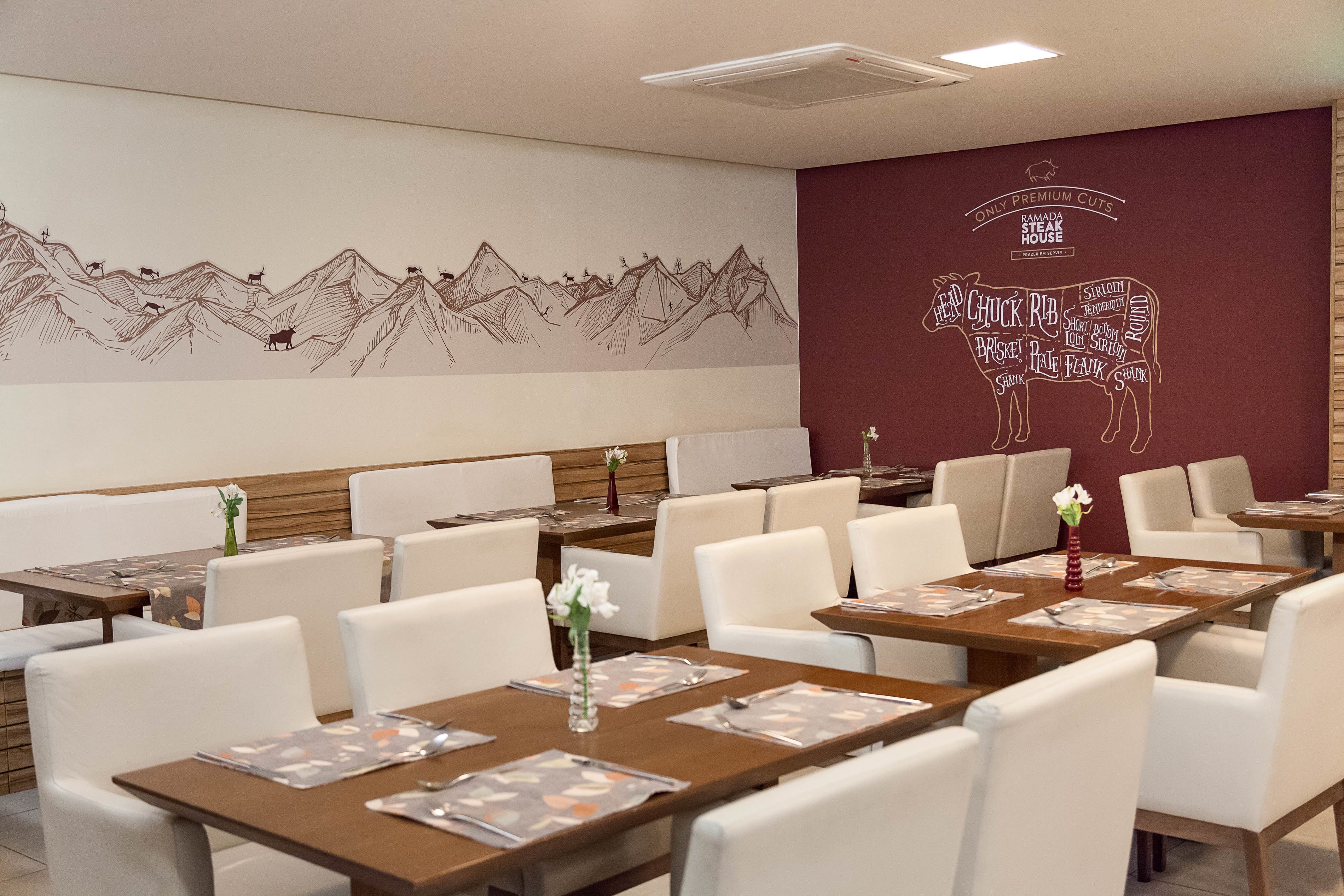 En restaurant med hvide stole og borde, et vægmaleri med bjerge og dyr, samt et skilt for "RAMADA STEAK HOUSE".