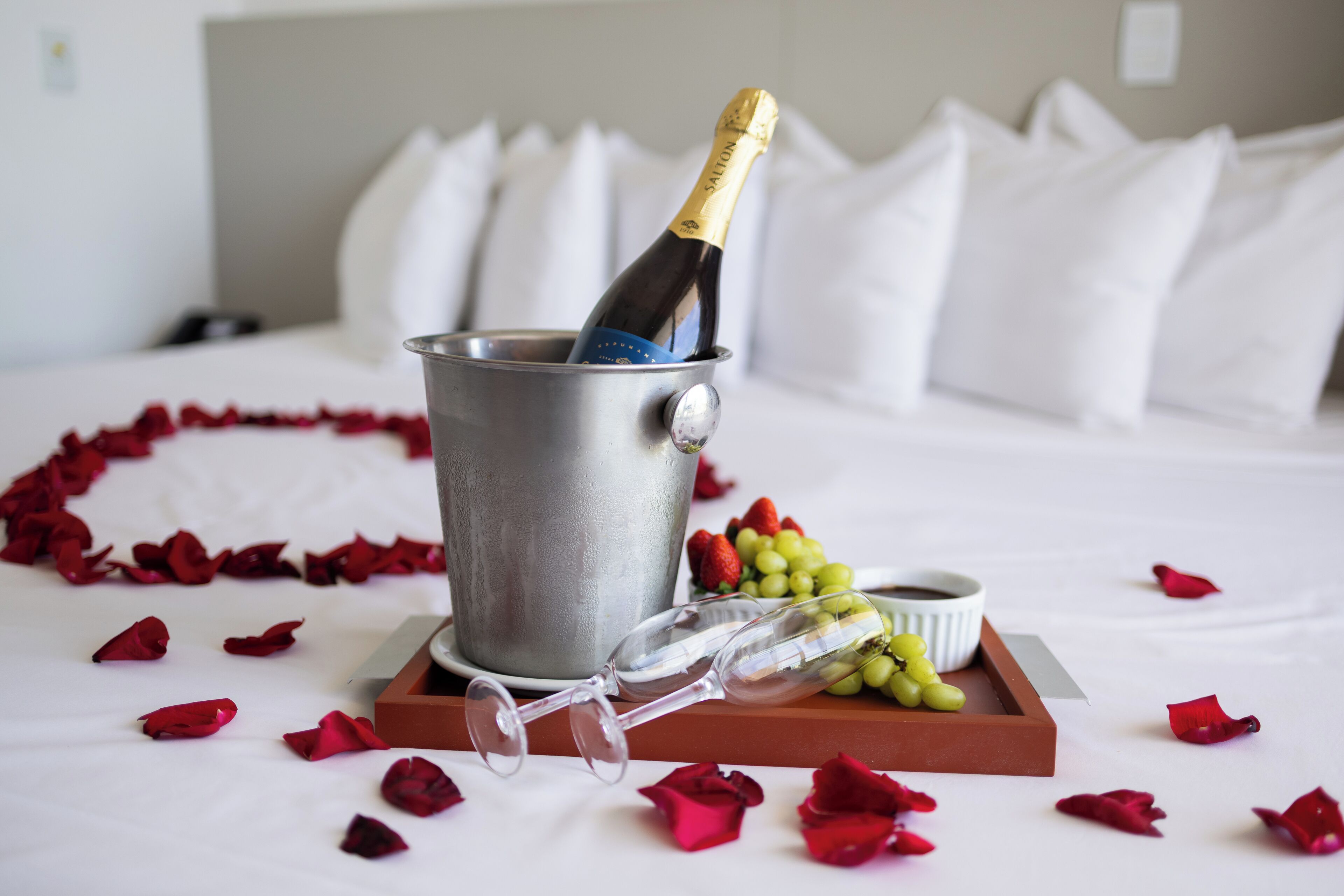 A bed with white bedding, a bucket with a bottle of champagne, a tray with wine glasses and grapes, and scattered rose petals.