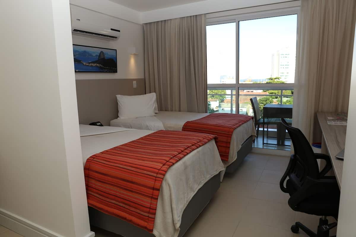 A hotel room with two beds, a desk, a chair, and a large window with a view of buildings and greenery.