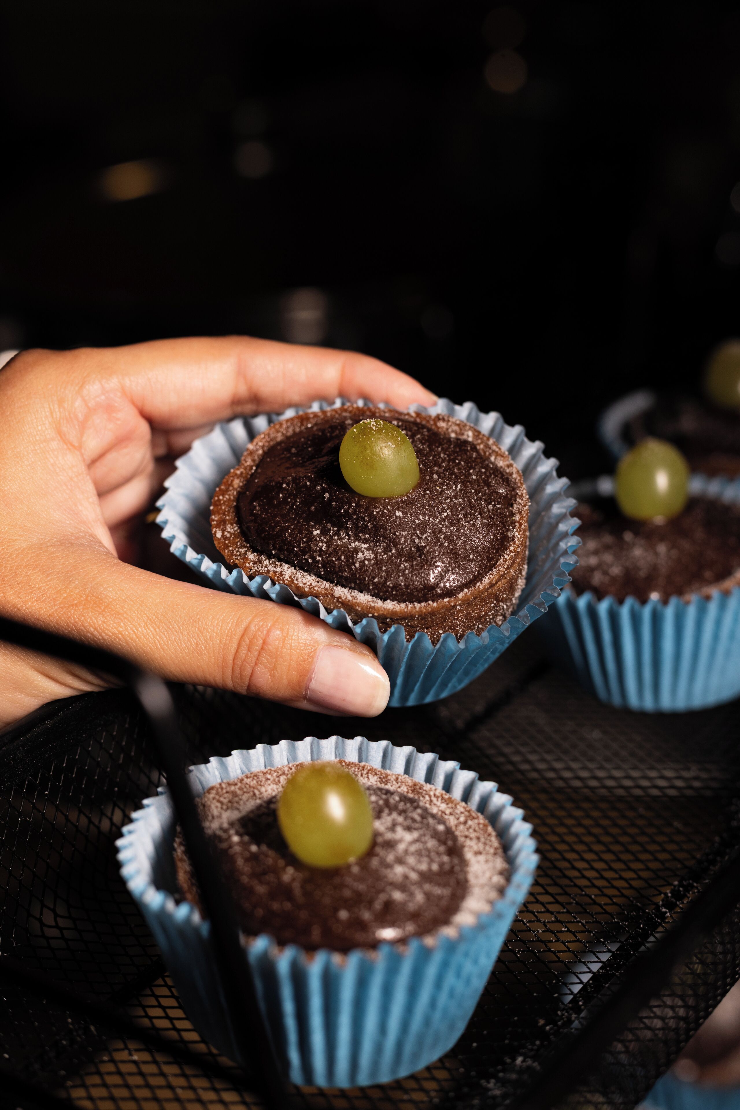 En hand håller i en chokladmuffin med en grön vindruva ovanpå.