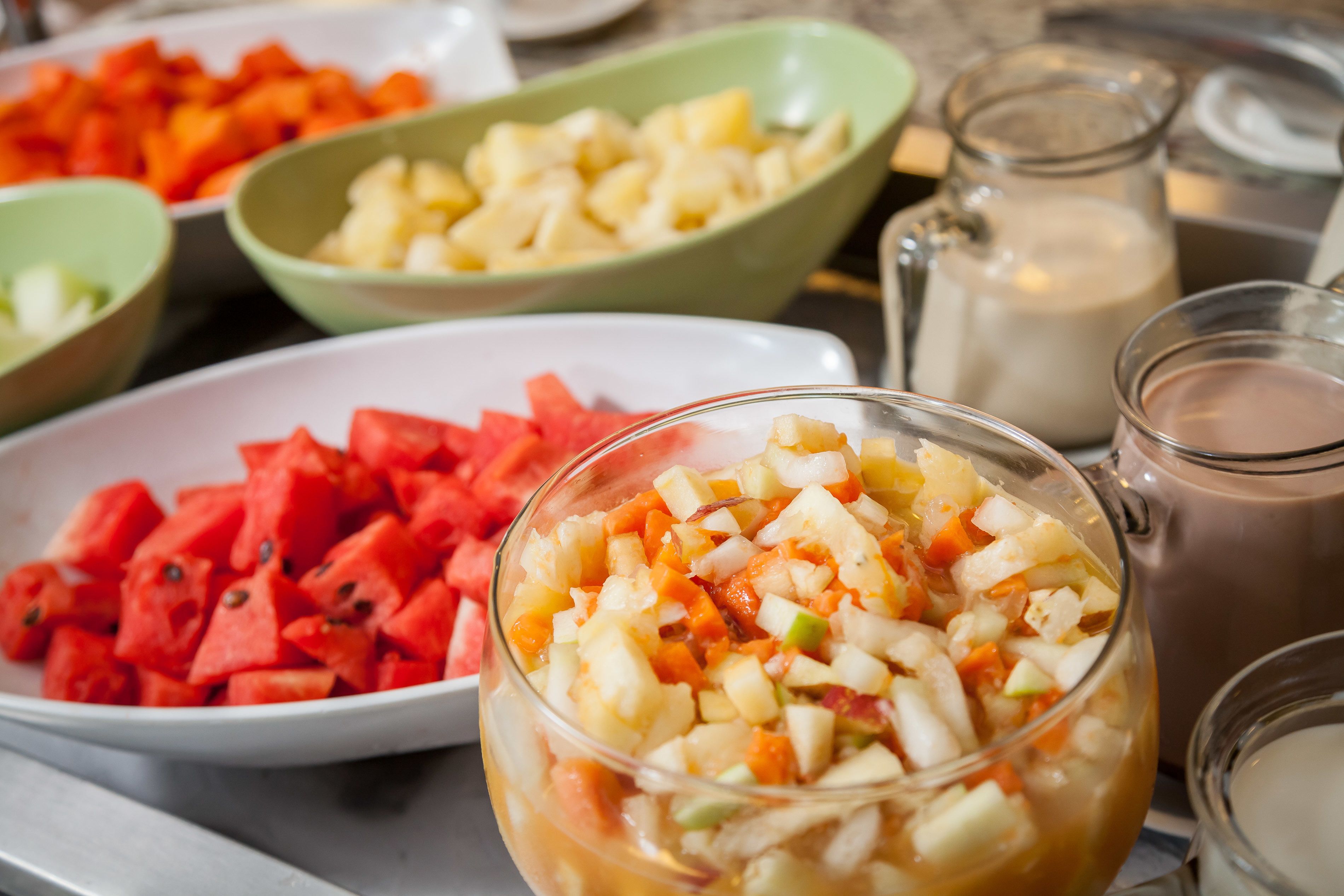 A table with various bowls of chopped fruits and a pitcher of milk.
