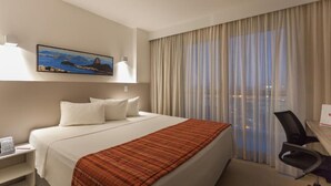 A hotel room with a bed, a desk with a chair, a painting on the wall, and a window with curtains. -  Transamerica Executive Macaé (Macaé)
