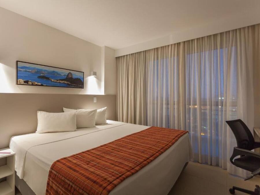 A hotel room with a bed, a desk with a chair, a painting on the wall, and a window with curtains.