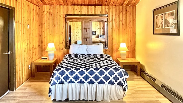 Un dormitorio con paredes de paneles de madera, una cama con colcha estampada, dos lámparas de noche, un espejo sobre la cama y un cuadro en la pared.