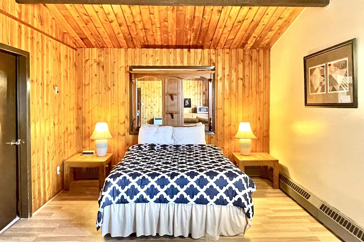 A bedroom with wooden panel walls, a bed with a patterned bedspread, two bedside lamps, a mirror above the bed, and a framed picture on the wall.